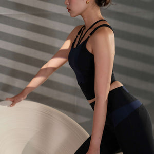 Yoga pose image featuring model wearing YOGASANA Cassie Cross-Back Performance Sports Bra in Misty London black and blue, arms stretched overhead while seated on a sculptural chair.