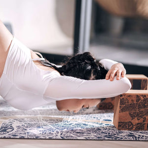 Woman practicing yoga on Yogasana Misty London map mat, stretching forward with cork yoga blocks beside her.