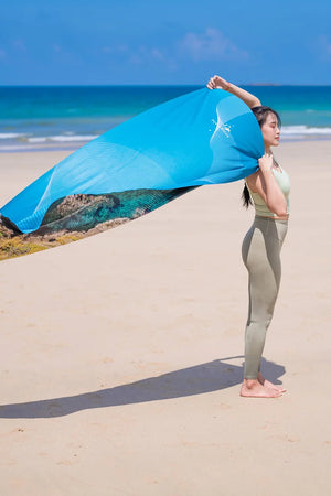 Yogasana Island Garden yoga towel lifted in the breeze by the ocean, featuring double-sided design in blue and earthy tones.