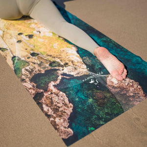 Yogasana Island Garden yoga towel detail shot on sandy beach, showcasing rocky shoreline print and vibrant turquoise tones.